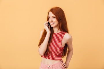 Happy  young beautiful redhead woman posing isolated over yellow background talking by mobile phone.