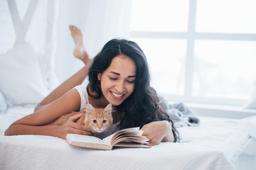 Home calm. Attractive blonde resting on the white bed with her cute kitten