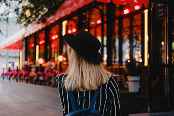 Obraz premium Blonde girl in black hat with backpack walking on the street in evening on background of red and yellow lights. City traveling alone concept