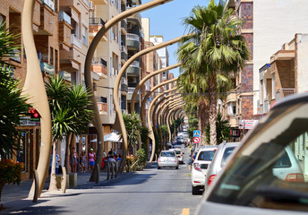 Busy Caballero de Rodas street Torrevieja resort town unusual bended street lights along the urban road in the center of city, lush palm trees walking people, Spain