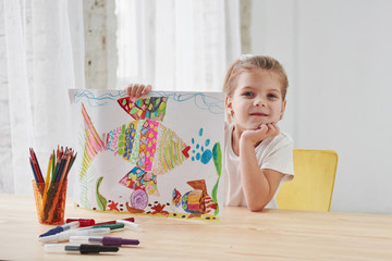 Nice daylight. Cute little girl in art school shows her first paintings drawn by pencils and markers