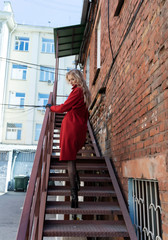 Fototapeta premium A beautiful blond girl wearing a red coat and gloves climbs a rusty metal staircase near a brick wall. Fashionable, commercial, advertising design. Copy space