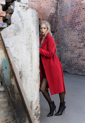 A beautiful blond girl wearing a red coat and gloves stands by a ruined wall and brick arch near an...
