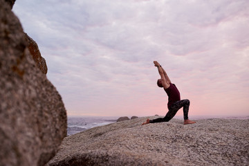 Man doing the cresent lunge pose on rocks at dusk