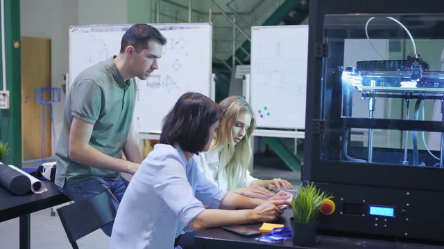 Team of three technicians using laptop computer to print prototype model in 3d printing machine in laboratory, panning shot