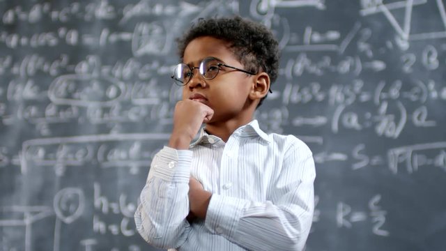Portrait shot of adorable little child prodigy in glasses standing before blackboard with complex mathematical formulas and thinking with hand on his chin