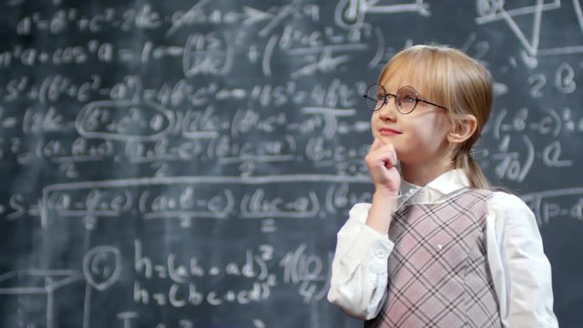 Portrait shot of cute little schoolgirl in glasses standing against blackboard with complex calculus formulas and thinking how to solve it