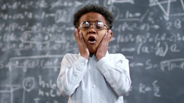 Portrait shot of cute little African-American genius boy in glasses standing before blackboard with complex math formulas and thinking, then exclaiming in surprise