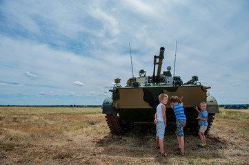 Fototapeta premium Three children in bright clothes are pushing away modern battle tank. Children play until tankists arrive. concept of contrasting innocent childhood and war