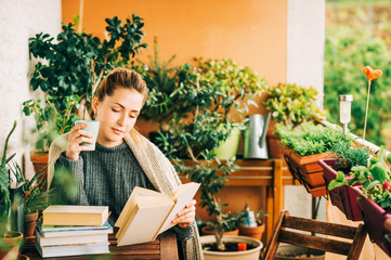 Young beautiful woman relaxing on cozy balcony, reading a book, wearing warm knitted pullover,...