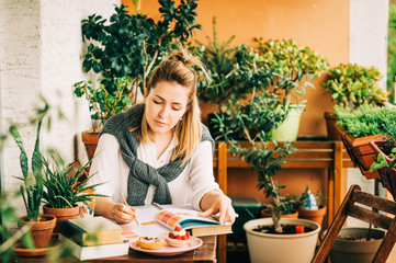 Young woman stuyding at home, sitting on the balcony, making notes in a nitebook