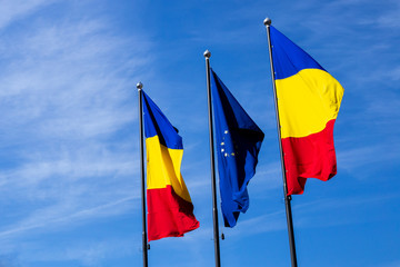 European Union emblem placed in the middle of two Romanian flags blowing in the wind on a bright blue sky in summer – Patriotic colors of Romania on metallic pole with copy space