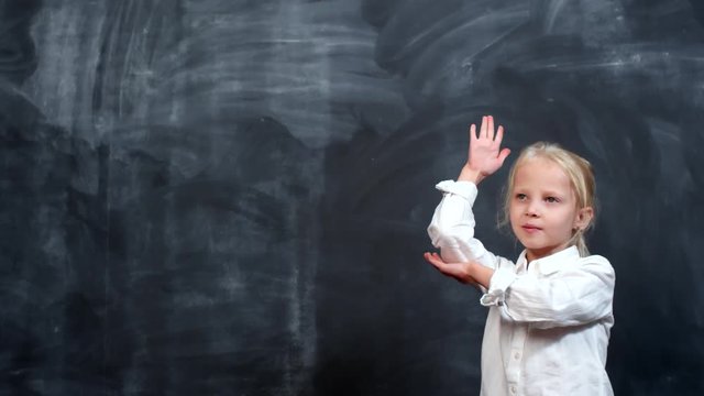 Portrait shot of cute little girl with blond hair standing against blackboard and raising her hand to answer question