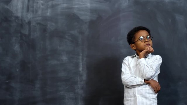 Portrait shot of cute little African-American boy in glasses standing against blackboard and thinking with his hand on chin