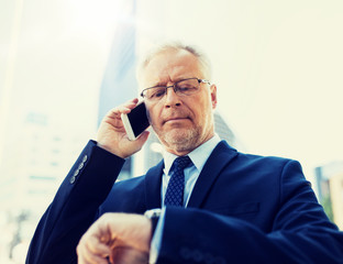 business, technology, time, punctuality and people concept - senior businessman calling on smartphone an looking at wristwatch or smart watch on his hand in city