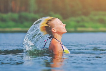 Active young blonde woman waving hair splashing water in river. Beautiful healthy lady relax and laughing, raising head out of the water. Vacation in paradise enjoying swimming. Motion freeze.