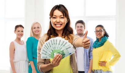 winning, saving and finances concept - happy asian young woman holding hundreds of euro money banknotes and showing thumbs up over group of people on background
