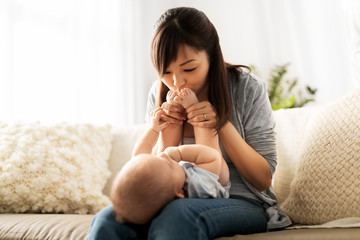 family and motherhood concept - happy smiling young asian mother kissing little baby son's feet at home