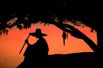 The silhouette of a man holding a sword sitting on a lake, a samurai and a river. During the sunset, men wear traditional Japanese clothes. - Powered by Adobe
