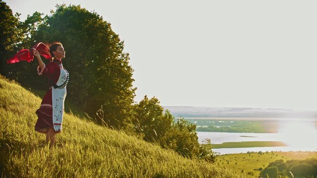 A young smiling woman in russian folk clothes standing on the field and enjoying the view - handkerchief flutters by the wind