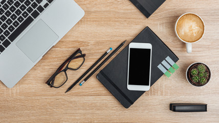 On the table in the office is a smartphone with a dark screen on a notepad, a laptop, glasses, stationery, a cactus and a cup of coffee. Top view. Moc kup.