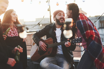 Just having fun together. Guy with acoustic guitar sings. Friends have fun at rooftop party with decorative colored light bulbs