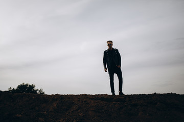 Young sexy man in leather jacket and sunglasses standing outdoor. Dark silhouette against grey sky