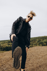 Young sexy man in leather jacket and sunglasses kicks in camera by leg outdoor