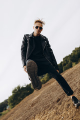 Young sexy man in leather jacket and sunglasses kicks in camera by leg outdoor
