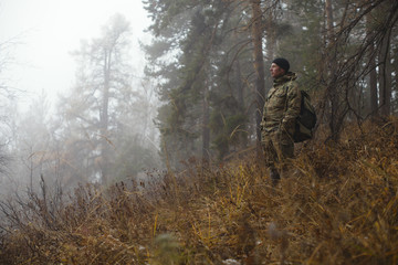 Traveler in camouflage clothes with a backpack observes the surroundings against the backdrop of a misty autumn forest in cold weather. Survival in the wild.