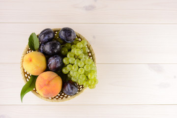 A bowl of tasty, ripe summer fruits on the white wooden table. In the bowl peach, plum and white grapes. Top view, copy space