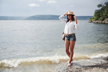A beautiful young girl in a hat and with a backpack playfully walks by the water. A warm summer day...