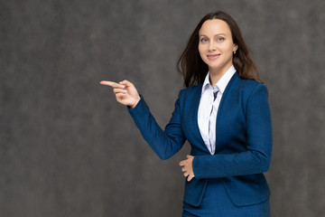 Portrait to the waist a young pretty brunette manager woman of 30 years in a business blue suit with beautiful dark hair. It is standing on a gray background, talking, showing hands, with emotions