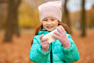 childhood, season and people concept - happy girl with smartphone at autumn park