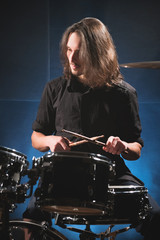 Fototapeta premium Portrait of a long-haired drummer with chopsticks in his hands sitting behind a drum set. Low key. Concepts of the creative freedom of the millenial generation