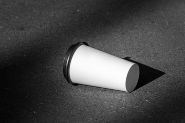 Close Up white paper cup with a black lid on the asphalt. Moke up. black and white photo. Disposable coffee cup