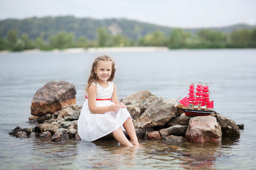 Little cute girl and scarlet sails. Girl sitting on the rocks on the seashore (ocean) with the ship. Happy childhood carefree game on the open sand, river lake, joy, fun. The concept of rest.