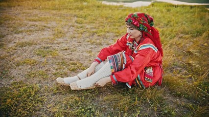 A woman in russian folk clothes putting on bast shoes - sitting on the field