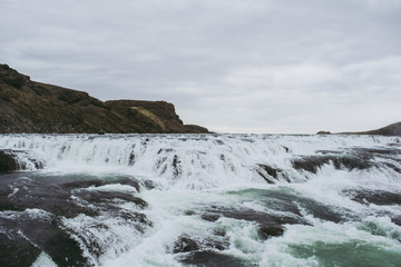 Cascade waterfall in Iceland. White splashes, turquoise water, and black rock under the gray sky. Spring