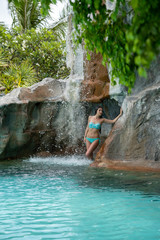 A young girl in a bathing suit standing under an artificial waterfall in the pool on site