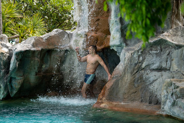 Fototapeta na wymiar A young guy stands under artificial waterfalls in the pool on site