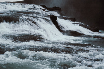 Cascade waterfall in Iceland. White splashes, turquoise water, and black rock under the gray sky. Spring