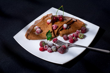 Pancakes on a white plate and a black background with juicy berries, condensed milk and powdered sugar.
