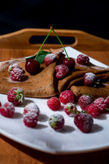  Pancakes on a white plate and a wooden tray with juicy berries, condensed milk and powdered sugar.