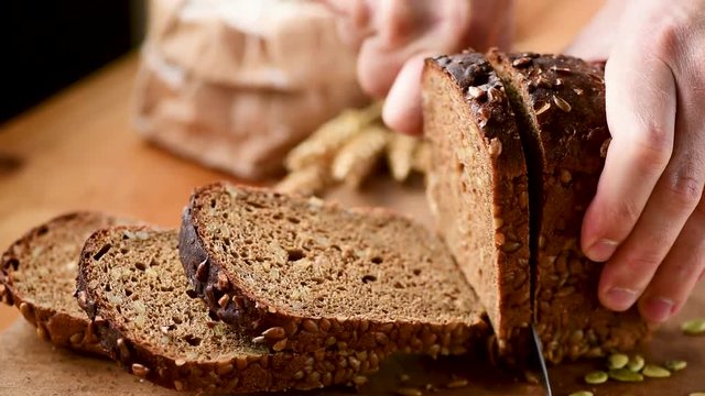 Person slicing fresh rye bread with seeds on cutting board. Closeup view