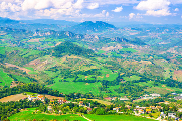 Fototapeta na wymiar Aerial top panoramic view of landscape with valley, green hills, fields and villages of Republic San Marino suburban district with blue sky white clouds background. View from San Marino fortress