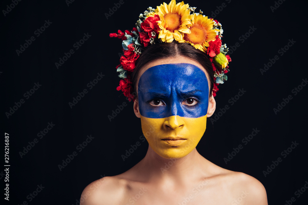 Wall mural sad naked young woman in floral wreath with painted Ukrainian flag on skin isolated on black