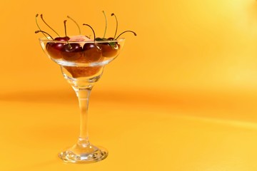 Glass transparent vase with a thin stem with ripe fresh peach and red sweet cherries with stalks. On a yellow background to the left.