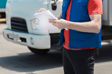 Courier driver in uniform making notes in document and delivery white truck behinde him