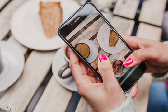 young unrecognizable woman taking a picture with mobile phone of her breakfast, a cup of coffee and a piece of cake. Breakfast concept